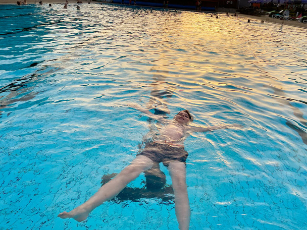 Школьный мальчик плескается в открытом бассейне в теплый летний день. Счастливый здоровый ребенок дошкольного возраста наслаждается солнечной погодой в городском общественном бассейне. Активность детей на открытом воздухе с водой. - Фото, изображение