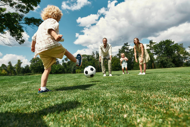 Little boy kicking the ball while parents and sister watching. Young family playing football on the grass field in the park on a summer day - Fotografie, Obrázek