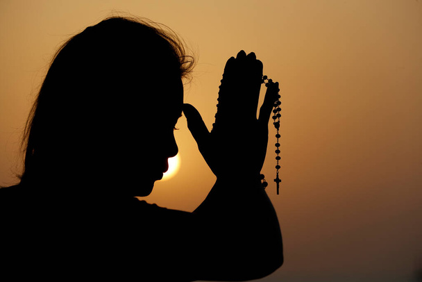 夕暮れ時にバラ色のビーズで祈る忠実な女性のシルエット。宗教、信仰、祈り、精神性の概念. - 写真・画像