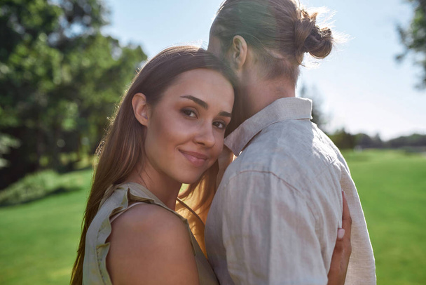 Portrait of pretty young woman smiling, embracing her husband, posing together outdoors while standing in the park - Фото, изображение