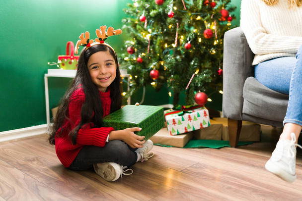 I feel so happy. Adorable latin girl sitting at home with her mom and opening presents during Christmas eve  - Foto, Imagen