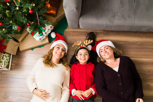 Blick von oben auf eine wunderschöne LGBTQ-Mutter mit gemischter Rasse, die es sich zu Hause mit ihrem entzückenden kleinen Mädchen zu Weihnachten gemütlich macht - Foto, Bild