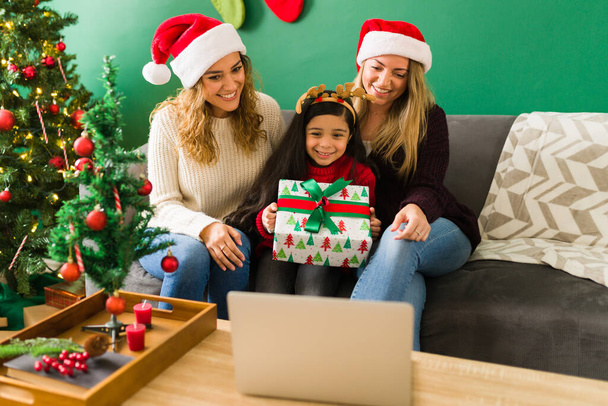 Un cadeau du Père Noël. Petite fille excitée montrant un beau cadeau à la famille lors d'un appel vidéo avec ses deux mères - Photo, image