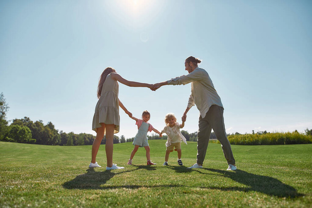 Happy young parents spending time together, holding hands in circle with their two little kids, boy and girl in green park on a summer day - 写真・画像
