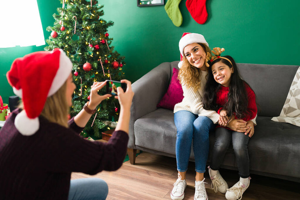 Mujer feliz tomando una foto con un teléfono inteligente a su esposa e hija caucásicas durante la víspera de Navidad  - Foto, Imagen