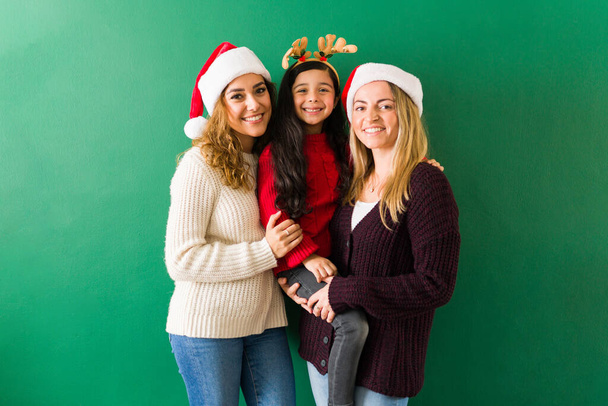 Noel Baba şapkalı LGBTQ çifti, sevimli kızlarını kucaklarken kameraya bakıyor. - Fotoğraf, Görsel
