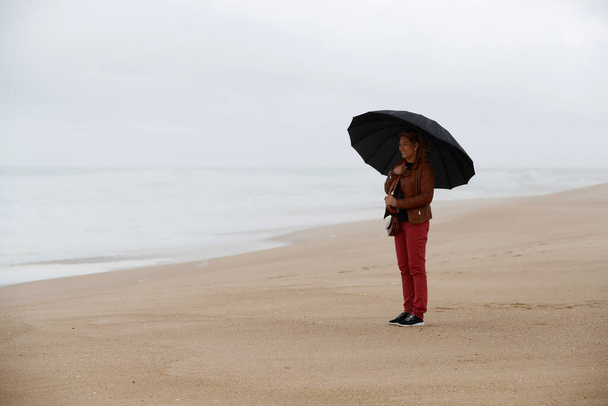 Женщина с черным зонтиком стоит перед морем в капризный и дождливый день. Назар. Португалия.  - Фото, изображение