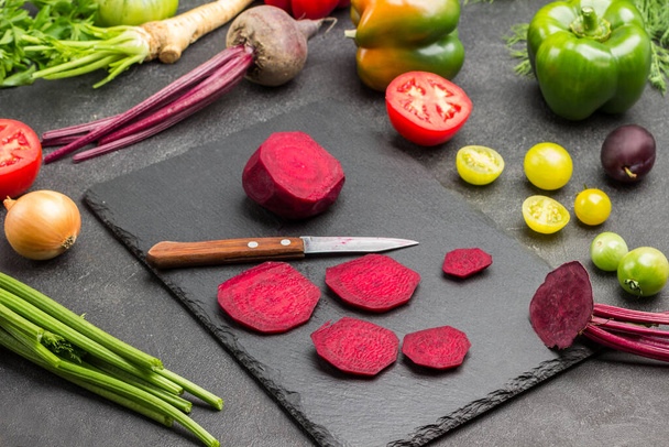 Chopped beets and kitchen knife on cutting board. Greens and vegetables on table. Black background. Top view - Foto, imagen