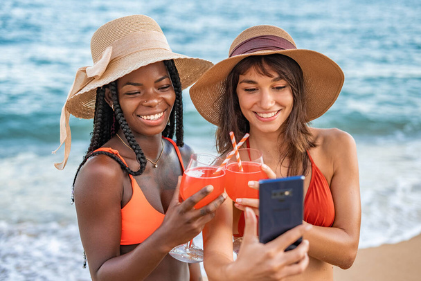 Γοητευμένες διαφορετικές φίλες με κοκτέιλ που παίρνουν selfie κοντά στη θάλασσα - Φωτογραφία, εικόνα