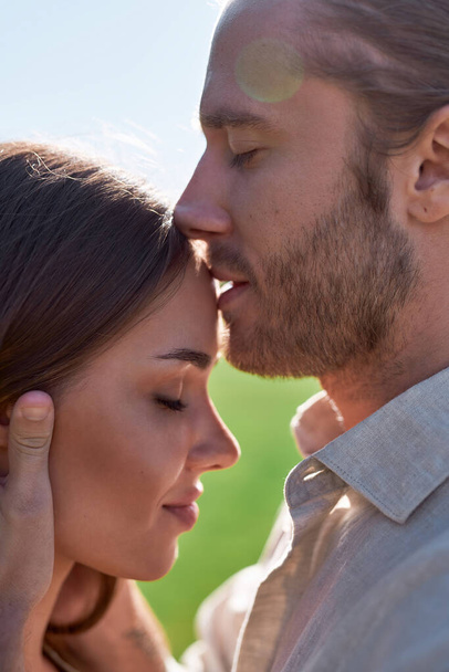 Close up portrait of sensuous young man kissing his wife in forehead while they posing together outdoors on summer day - Photo, image