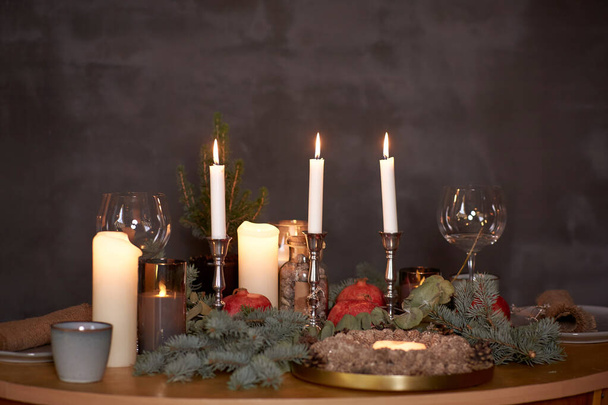 Elegant Christmas table setting with cozy candles, plates, pomegranate and branch of fir tree close up. Vintage grey wall copy space. Cozy home concept. High quality photo - Foto, Imagen