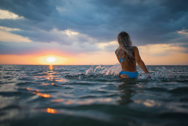 A girl with blond hair in a blue swimsuit splashes to the sides while sitting in an estuary on a sunset background - Foto, immagini