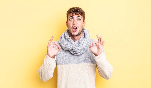 young handsome man feeling shocked, amazed and surprised, showing approval making okay sign with both hands - Fotoğraf, Görsel