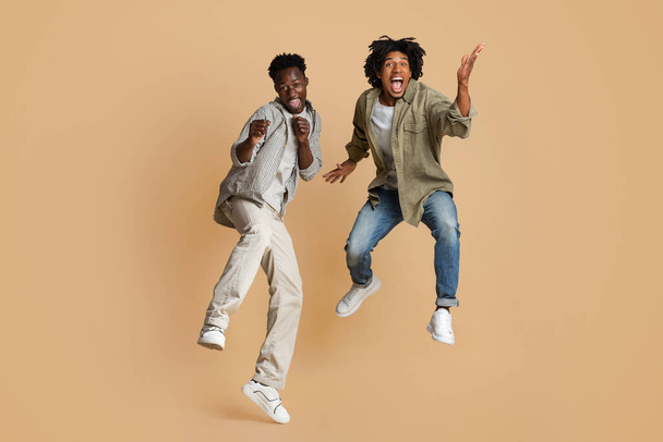 Τρελή διάθεση. Δύο ενθουσιασμένοι μαύροι που πηδούν πάνω από το φόντο του στούντιο Μπεζ - Φωτογραφία, εικόνα