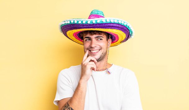 Młody, przystojny mężczyzna uśmiecha się ze szczęśliwą, pewną siebie miną z ręką na podbródku. meksykańska koncepcja kapelusza - Zdjęcie, obraz