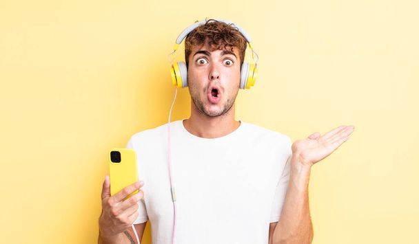 young handsome man looking surprised and shocked, with jaw dropped holding an object. headphones and smartphone concept - Foto, afbeelding