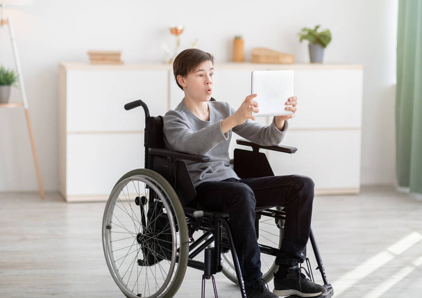 Järkyttynyt vammainen teini-ikäinen poika pyörätuolissa käyttäen tablet-tietokonetta kotona, selaamalla Internetiä tai katsomalla siistiä videota verkossa - Valokuva, kuva