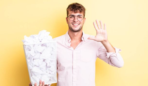 young handsome man smiling and looking friendly, showing number five. paper balls trash concept - Fotoğraf, Görsel