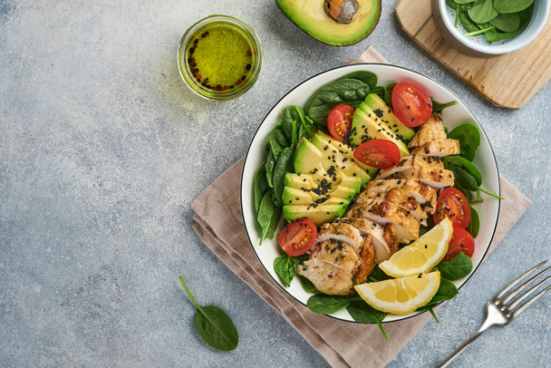 Fresh green salad with grilled chicken fillet, spinach, tomatoes, avocado, lemon and black sesame seeds, olive oil in white bowl on light slate background. Nutrition Diet Concept. Top view. copy space - Foto, Bild