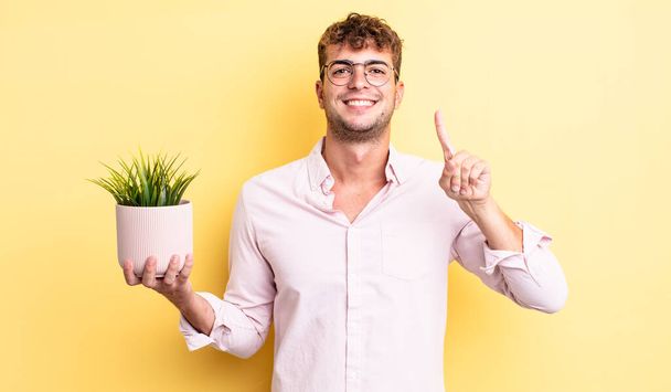 young handsome man smiling and looking friendly, showing number one. decorative plant concept - Foto, Imagem