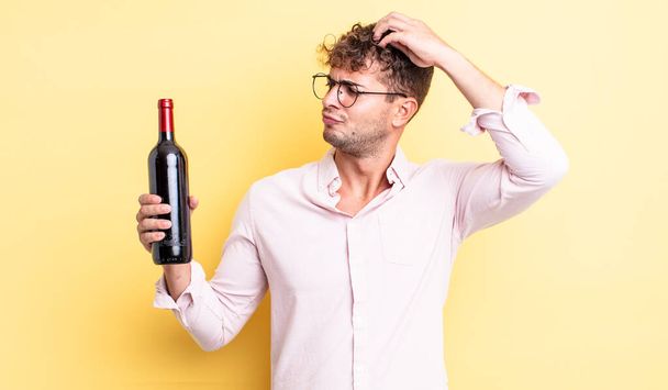 young handsome man feeling puzzled and confused, scratching head. wine bottle concept - Foto, immagini
