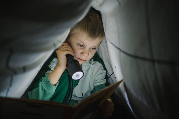 Little boy reading a book at night with lantern covered with blanket - Foto, imagen