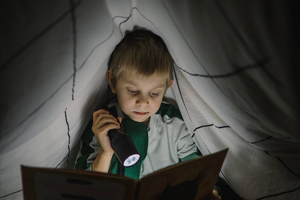 Little boy reading a book at night with lantern covered with blanket - Фото, зображення