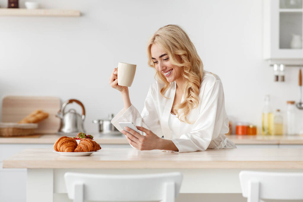 Souriant jeune jolie dame blonde caucasienne en peignoir prendre le petit déjeuner, boissons café et dactylographier sur smartphone - Photo, image