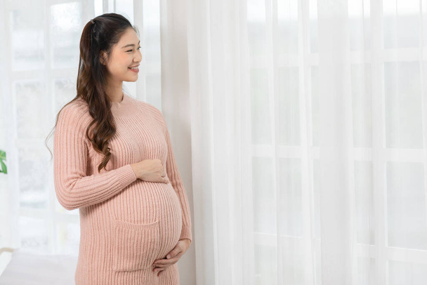Happy Pregnant Woman standing in front of windows and stroking her big belly with love at cozy home,Pregnancy of young woman enjoy with future life relax with love,Motherhood and Pregnant Concept - Zdjęcie, obraz