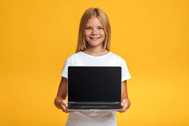 Vidám serdülő szőke lány tanuló show laptop üres képernyőn, elszigetelt sárga alapon - Fotó, kép
