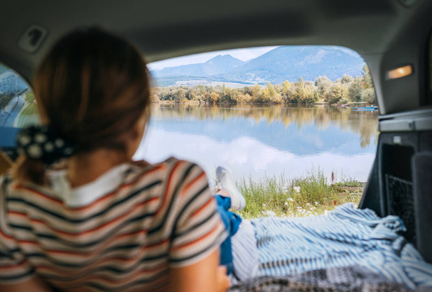 Woman lying in the cozy car trunk and enjoying the mountain lake view in the morning after the camper night. Warm early autumn auto traveling concept image.   - Valokuva, kuva