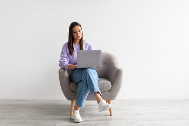 Hymyilevä nuori valkoihoinen nainen istuu nojatuolilla kannettavan tietokoneen kanssa valkoista studioseinää vasten, kopioi tilaa - Valokuva, kuva