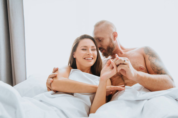 Una pareja enamorada yace en la cama bajo las sábanas. La mujer se ríe, y el hombre toca suavemente su cabeza con su nariz - Foto, imagen