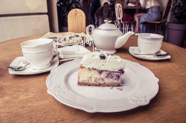 白い磁器プレート上のブルーベリーチーズケーキ。街のカフェのテーブルでティーポットやカップの静物画. - 写真・画像