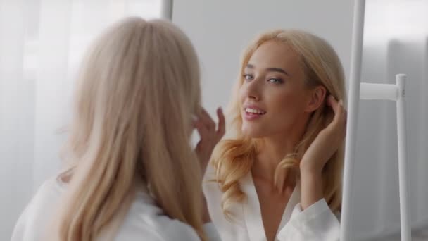 Nuori kaunis onnellinen nainen katsoo peiliin, koskettaa kasvojaan ja hymyilee - Materiaali, video