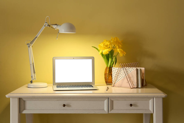 Local de trabalho elegante com lâmpada brilhante e laptop perto da parede de cor à noite - Foto, Imagem
