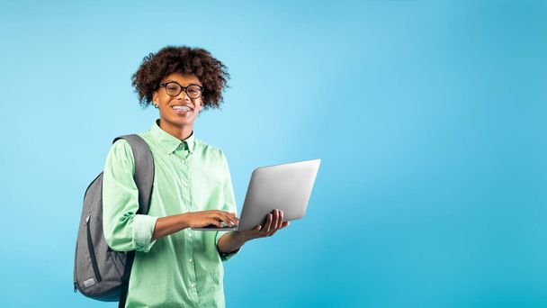 Koncepcja technologii i e-learningu. Podekscytowany czarny nastolatek facet z laptopem i plecakiem, niebieskie tło, puste miejsce - Zdjęcie, obraz