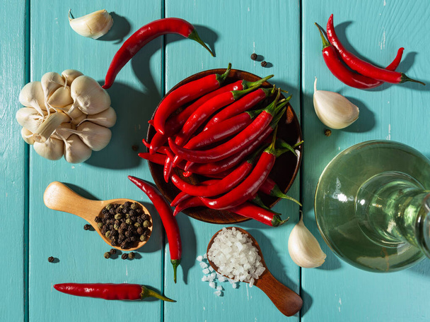 Directly above view of raw red hot chili pepper pods, sea salt and whole black peppercorns in wooden spoons, garlic head and olive oil glass jar cast shadows in a bright light. Spices and seasonings. - Valokuva, kuva