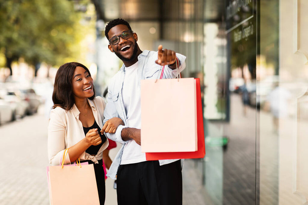 Веселая африканская пара с пакетами для покупок, указывающими на окно - Фото, изображение