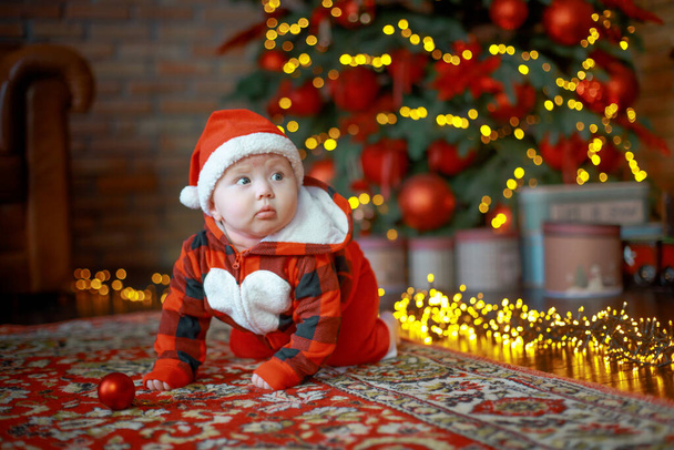 Klein gelukkig meisje van 6 maanden kruipt in de buurt van een nieuwjaarsboom op kerstavond. kind in Santa Claus kostuum in de buurt van lichtgevende bloemenslingers. Gelukkig nieuwjaar 2020 concept - Foto, afbeelding