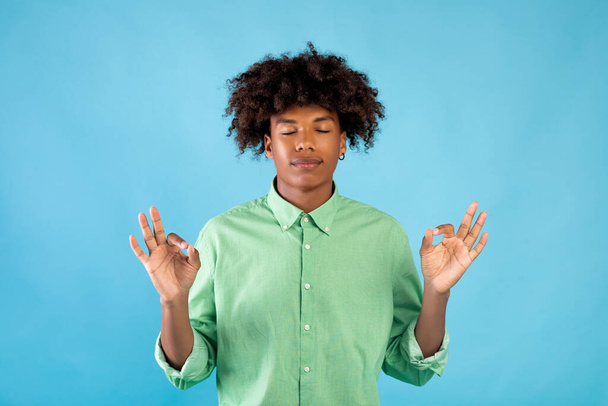 Tranquilo chico afroamericano meditando, practicando yoga, ejercicios de respiración con los ojos cerrados, fondo azul - Foto, imagen