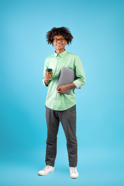 Modern diák. Boldog fekete srác laptop számítógép és elvitelre kávé pózol kamera kék háttér - Fotó, kép