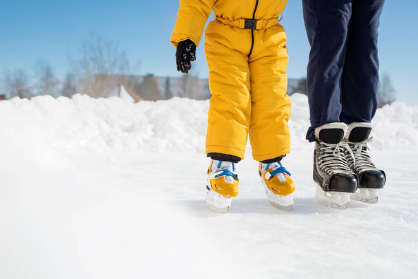 Ein Mädchen in Overalls lernt an einem sonnigen Tag mit ihrem Vater Schlittschuhlaufen auf der städtischen Eisbahn. Der Mann stützt das Kind an der Hand. Eiskunstlauf-Lehrer. EA-Eislaufkurse. Ohne Gesicht - Foto, Bild