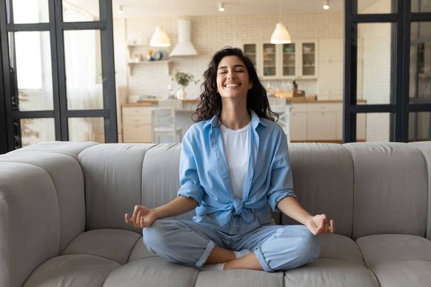 Cheerful young lady sitting on comfy couch, meditating with closed eyes at home, full length - Foto, Imagen