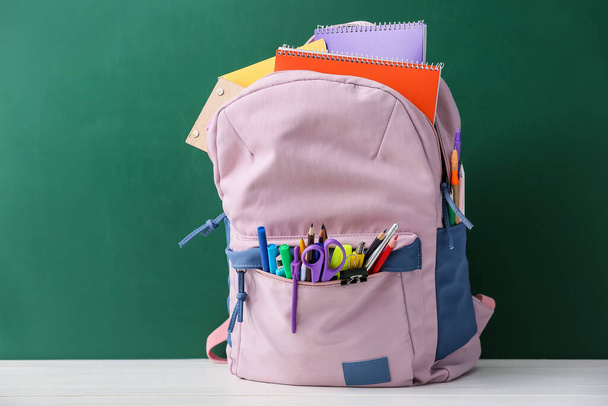 Κομψή σχολική τσάντα πλάτης με γραφική ύλη στο τραπέζι εναντίον μαυροπίνακα - Φωτογραφία, εικόνα
