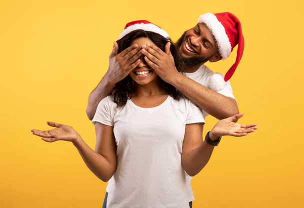 Empolgado homem afro-americano fechando os olhos de esposas, surpreso jovem sorrindo, casal vestindo chapéus Sants - Foto, Imagem