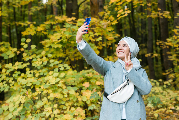 Muchacha asiática musulmana joven de moda en hijab tomando una selfie en el teléfono inteligente al aire libre en el parque de otoño. Espacio de copia. - Foto, Imagen