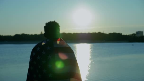 Afroamerikaner mit amerikanischer Flagge auf den Schultern blickt bei Sonnenaufgang in die Ferne - Filmmaterial, Video