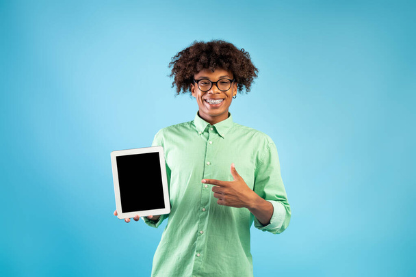 Szczęśliwy czarny nastolatek wskazując na tablet z makietą na stronie internetowej lub aplikacji na ekranie, niebieskie tło studio - Zdjęcie, obraz