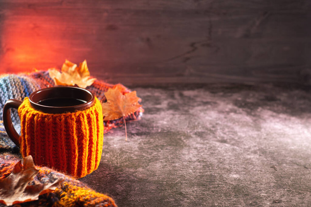 Filiżanka kawy w dzianinowej kurtce na stole z kolorowym dzianinowym szalikiem, liśćmi klonu. Jesienny skład sezonowy. Pomarańczowe podświetlenie, drewniane czarne tło - Zdjęcie, obraz